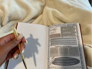 Hand holding a gold pen over an open Bible study journal on a cozy blanket